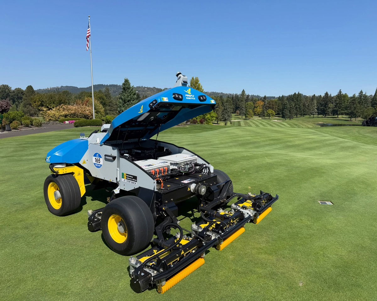 firefly robotics fairway reel mower with hood up to show battery and control boxes inside, sitting on green grass with striped golf course fairways in the background